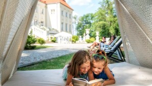 Twee kinderen lezen een boek op een witte buitensauna met uitzicht op een gebouw en groen in de verte.