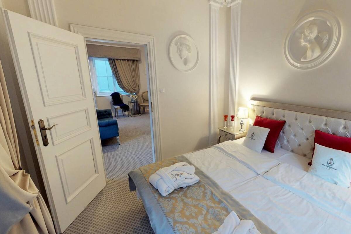 Une chambre d’hôtel avec un grand lit, deux oreillers rouges, une tête de lit blanche et une petite table avec une lampe et des verres.