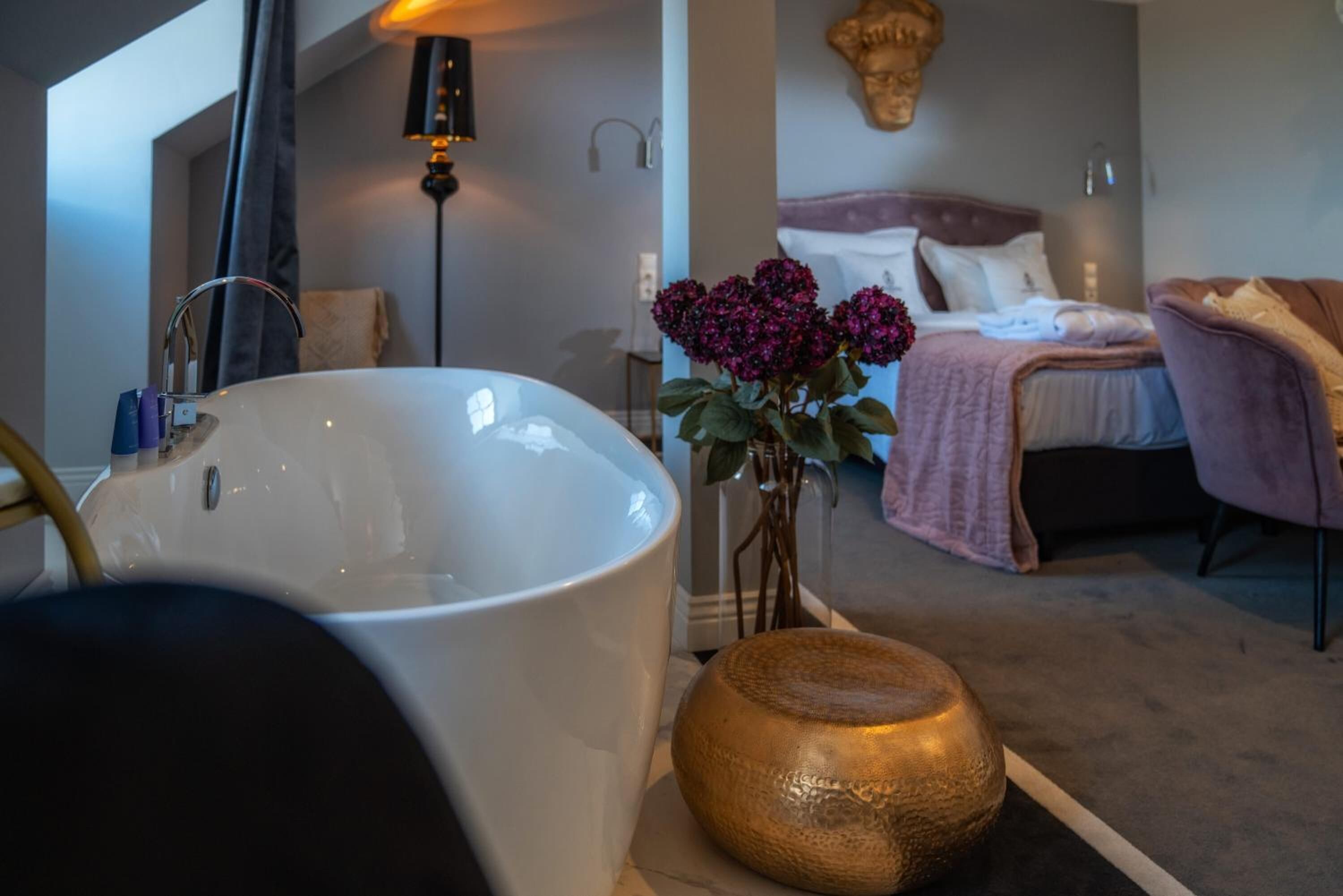 A bathroom with a large white bathtub, a golden decorative bowl, and a vase with purple flowers.
