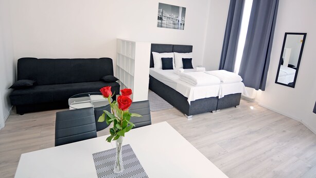 Une chambre d’hôtel moderne avec un canapé noir, un lit avec une literie blanche, une petite table sur laquelle est posé un vase de roses rouges, et un miroir au mur.