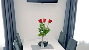 Une table à manger blanche avec quatre chaises, un téléviseur à écran plat fixé au mur et un vase rempli de roses rouges posé sur la table.