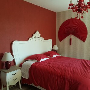 Un dormitorio con cama blanca, ropa de cama roja, un ventilador rojo y un candelabro rojo.