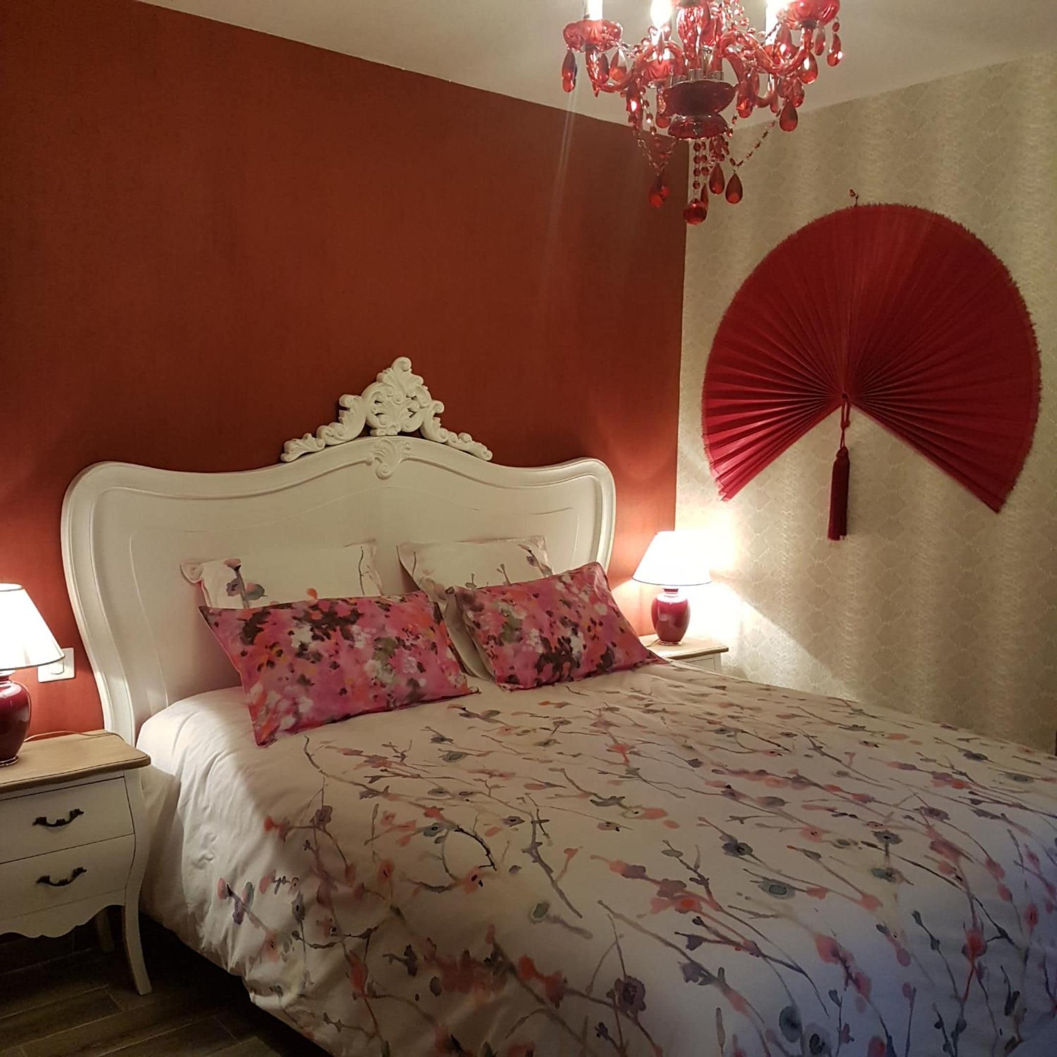 A bedroom with a white headboard, floral bedding, a red fan, and a chandelier.