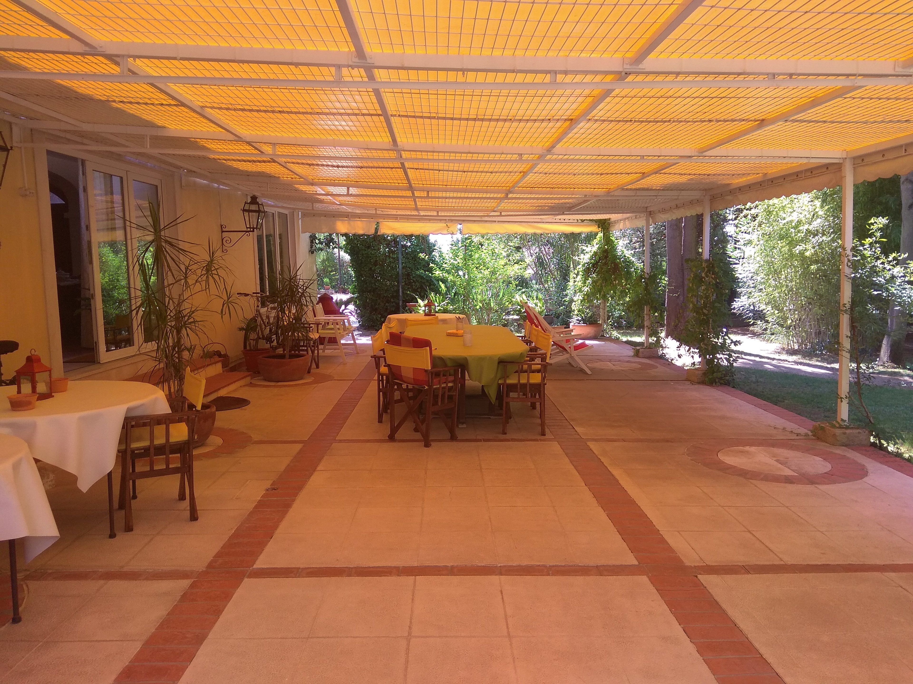 Un espace extérieur aménagé en terrasse, avec des tables et des chaises, recouvert d’un auvent jaune et entouré de verdure.