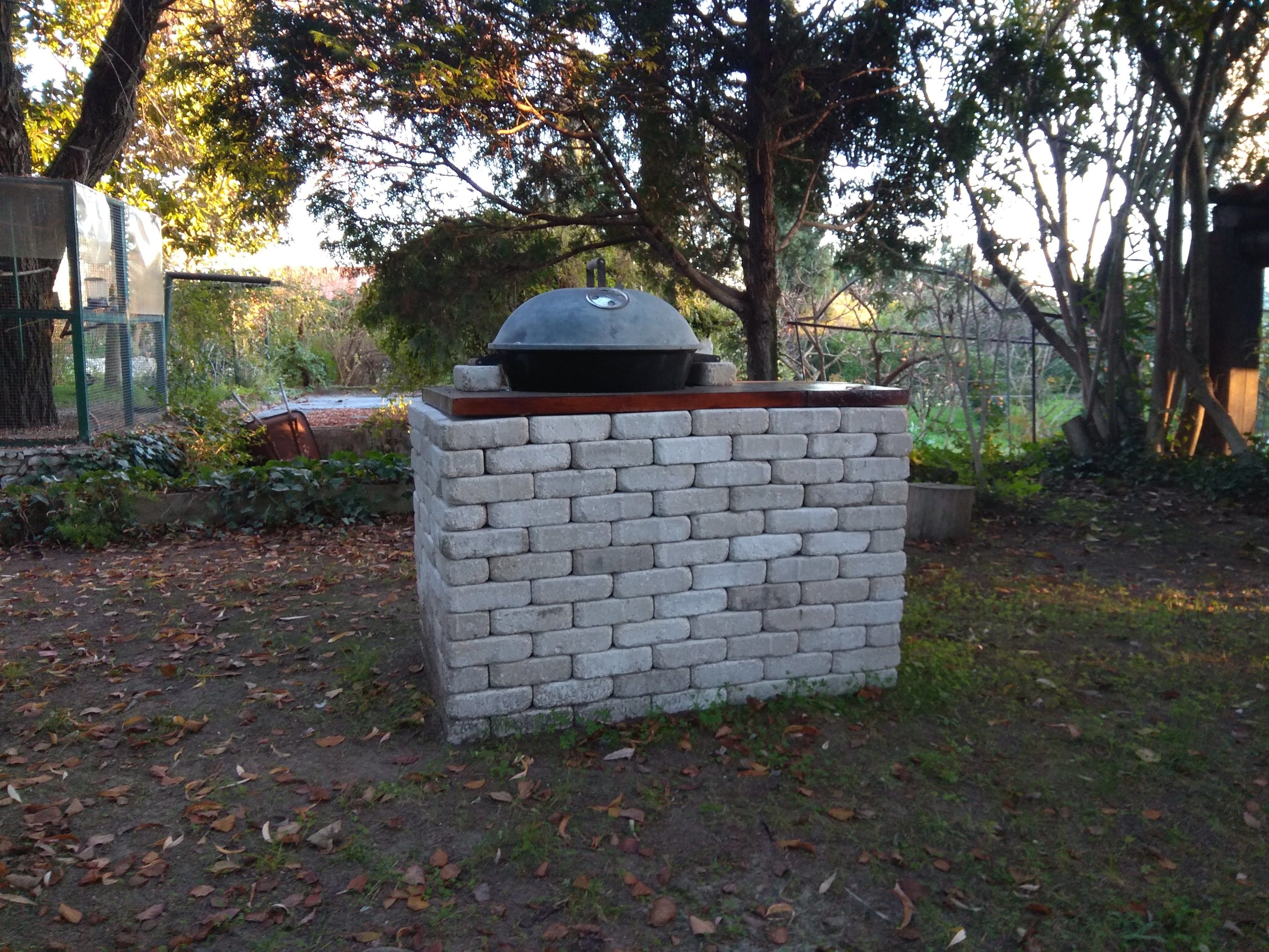 Una parrilla de ladrillo con chimenea, rodeada por un jardín con árboles y una cerca.