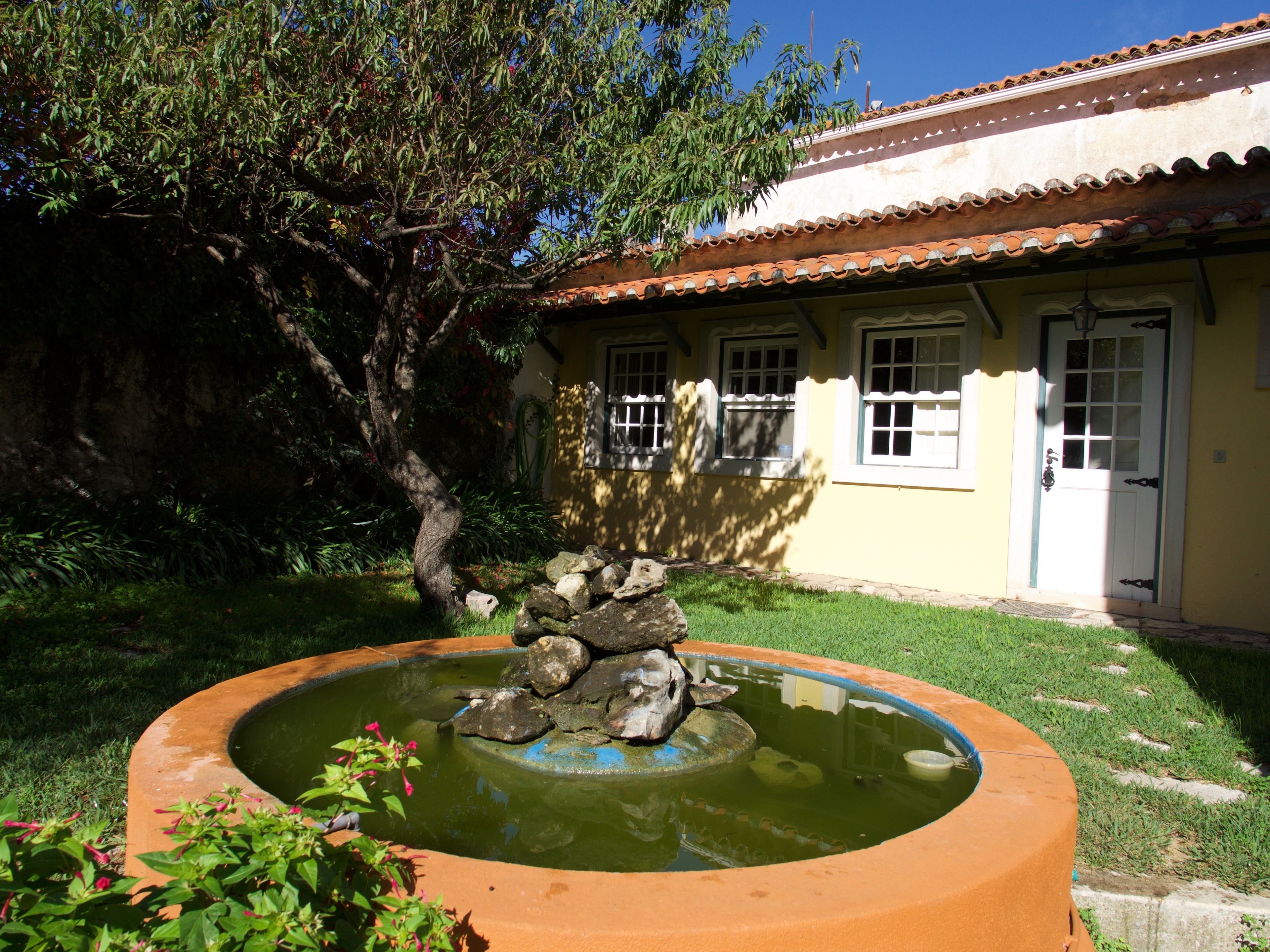Un jardín con un estanque circular, un árbol y un edificio con techo de tejas y una puerta.