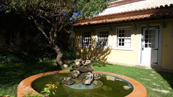 Un jardín con un estanque circular, un árbol y un edificio con techo de tejas y una puerta.