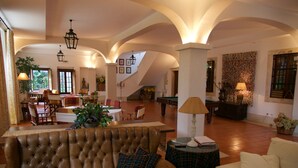 Un vaste espace de vie comprenant un canapé en cuir marron, un canapé blanc et une table ronde avec une lampe. On y trouve également plusieurs chaises, une table de billard et un escalier menant à un niveau supérieur.