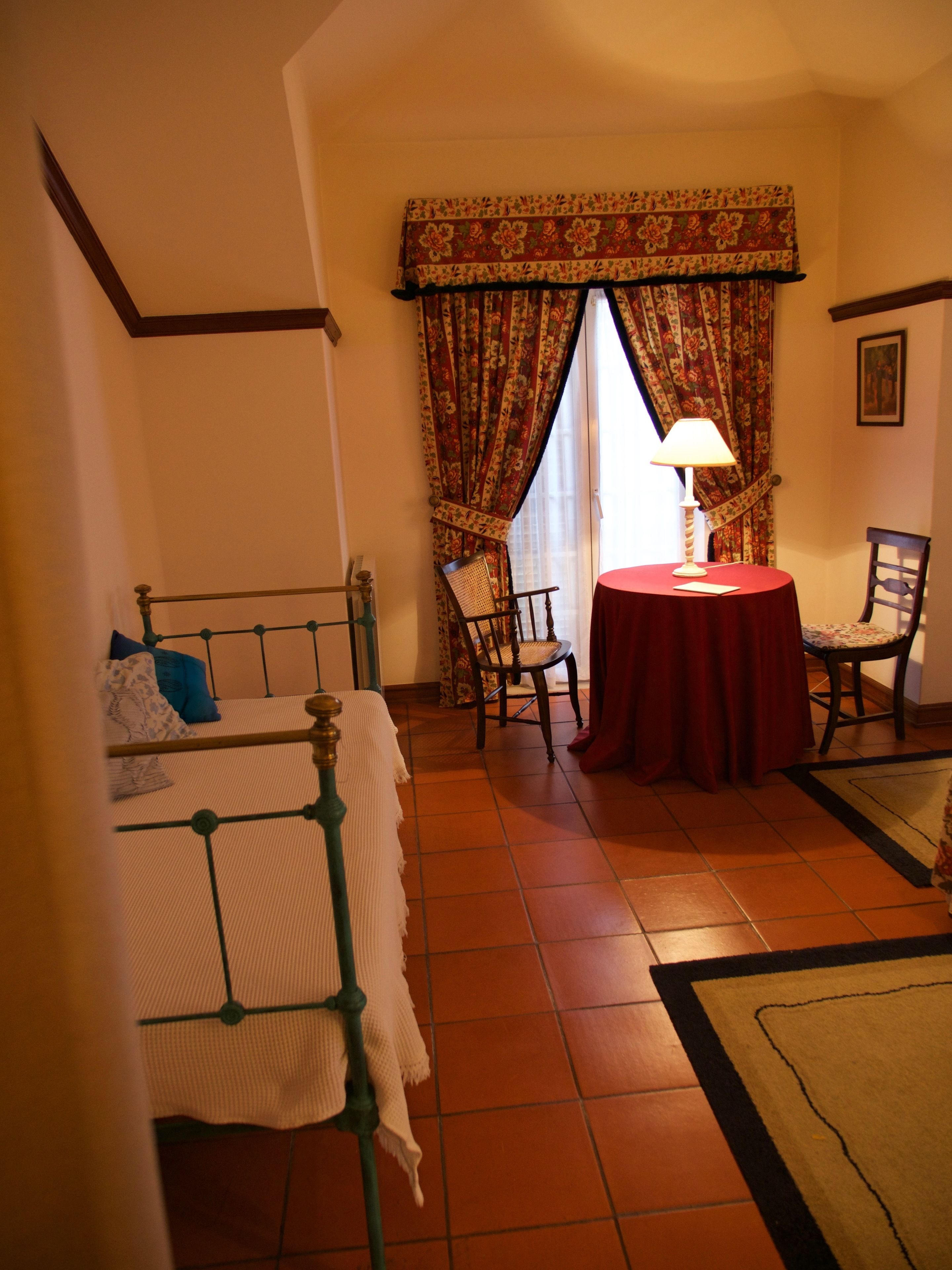 A bedroom with a bed, a chair, a table with a lamp, and a window with curtains.