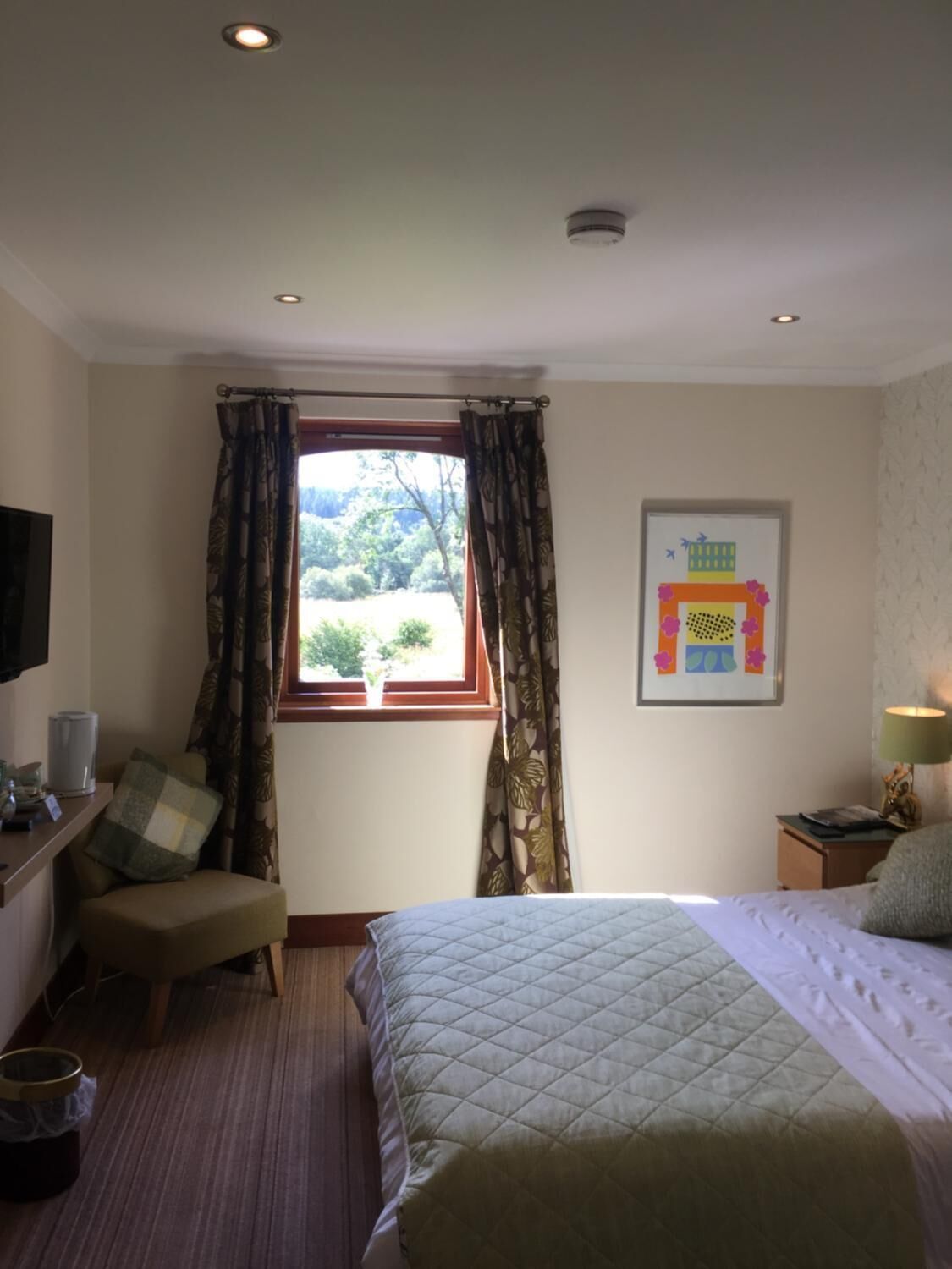 Habitación de hotel con cama, una silla, escritorio con lámpara, ventana con cortinas y un cuadro en la pared.