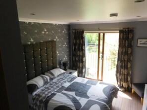 A bedroom with a large bed, a headboard with lights, a nightstand, and a view of the outdoors through a sliding door.