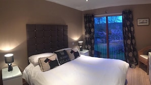 Habitación de hotel con una cama grande, dos lámparas de noche, una silla, un tocador y un balcón con vistas a árboles.