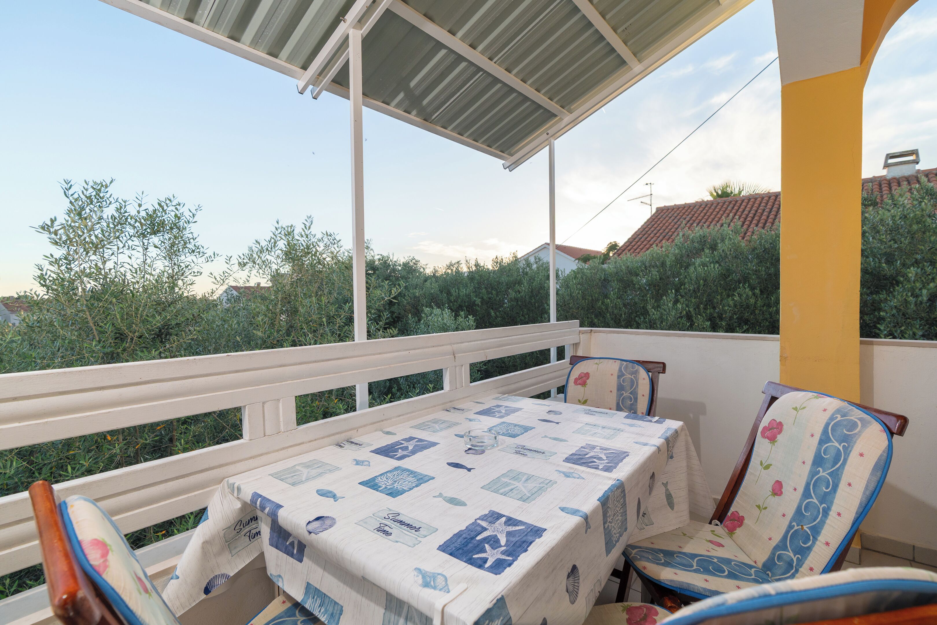 A covered balcony with a table and chairs, a white railing, and a view of greenery and buildings.