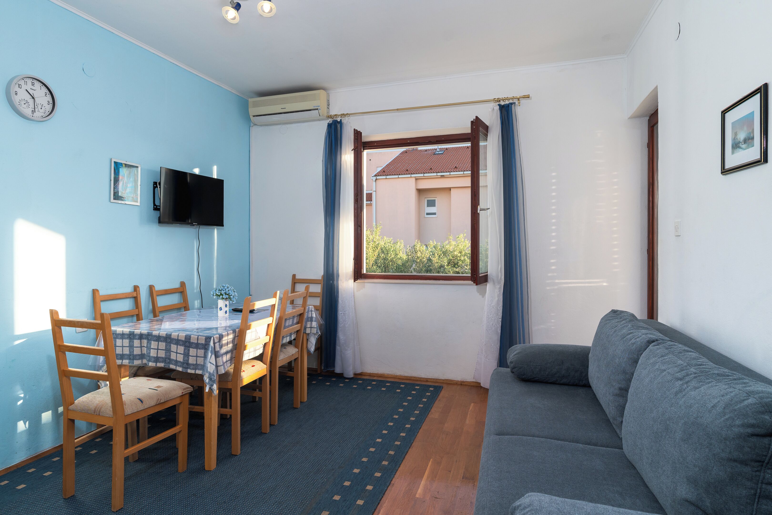 A room with a dining table, chairs, a grey sofa, a TV, and a window with curtains.