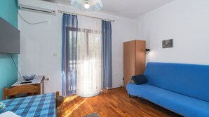 Una habitación con un sofá azul, suelo de madera, una mesa con mantel de cuadros, una silla azul, un mueble de madera y una lámpara en el techo.