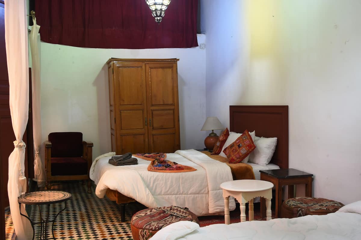 A bedroom with a bed, a wooden wardrobe, a chair, a table, and a patterned ottoman.
