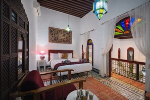 Un dormitorio con cama, mesita de noche, silla, mesa con tetera, lámpara colgante, ventana con vitrales y balcón con barandilla.
