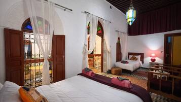 A hotel room with a large bed, a smaller bed, a sitting area with a table, and a view through a window with a balcony.