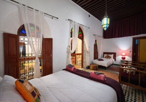 Habitación de hotel con una cama grande, otra más pequeña, un rincón de descanso con una mesa y vistas a través de una ventana con balcón.