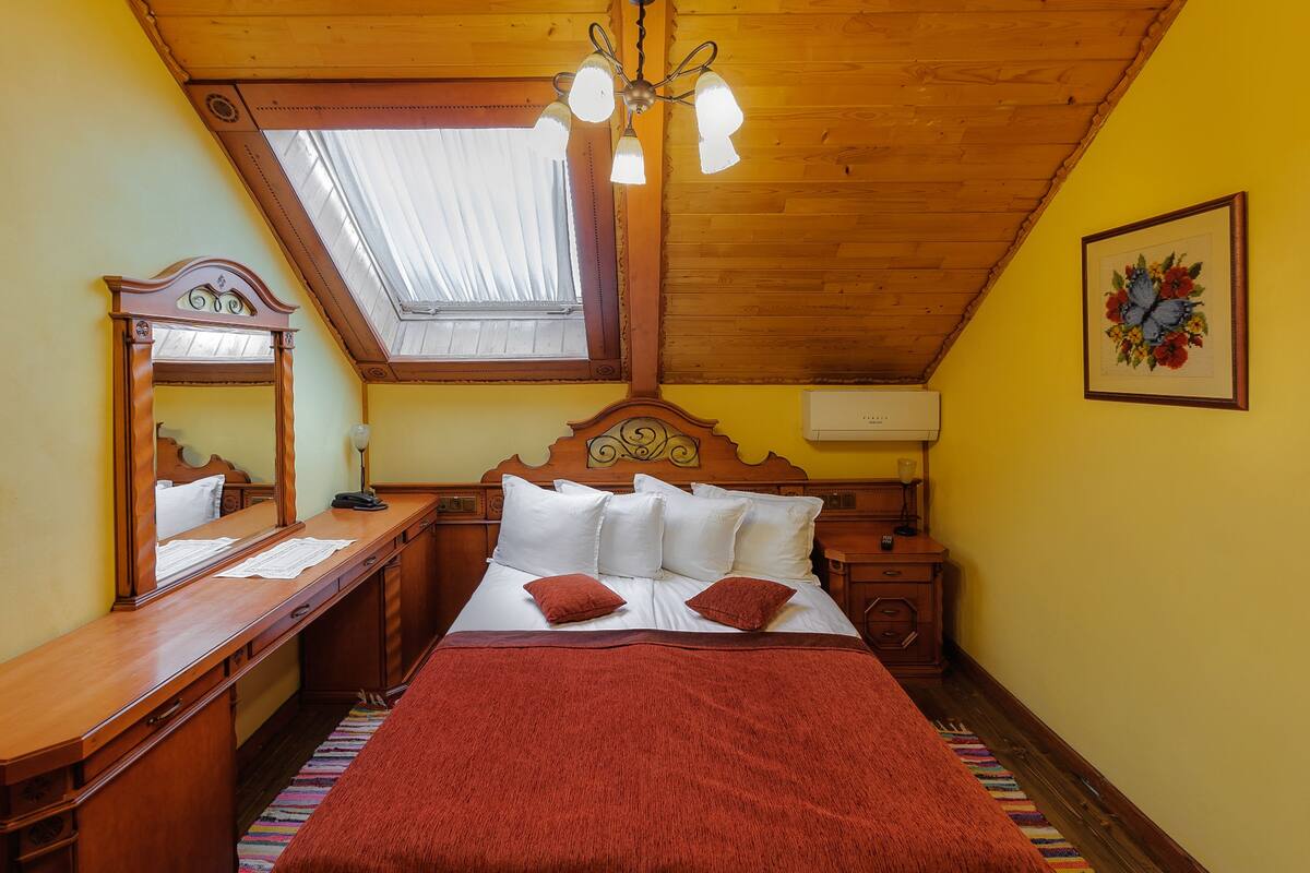 A bedroom with a sloped ceiling, a wooden headboard, a large bed with a red blanket, a nightstand, a bedside table, a mirror, and a window with a skylight.