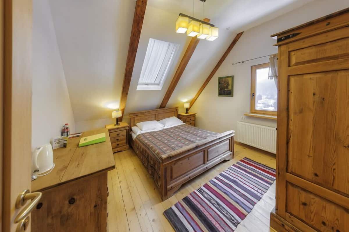 A bedroom with a wooden bed, a nightstand, a dresser, a window with a view, and a sloped ceiling with exposed beams.