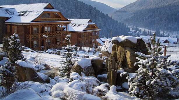 A wooden lodge with a snow-covered roof, surrounded by snow-covered trees and mountains.