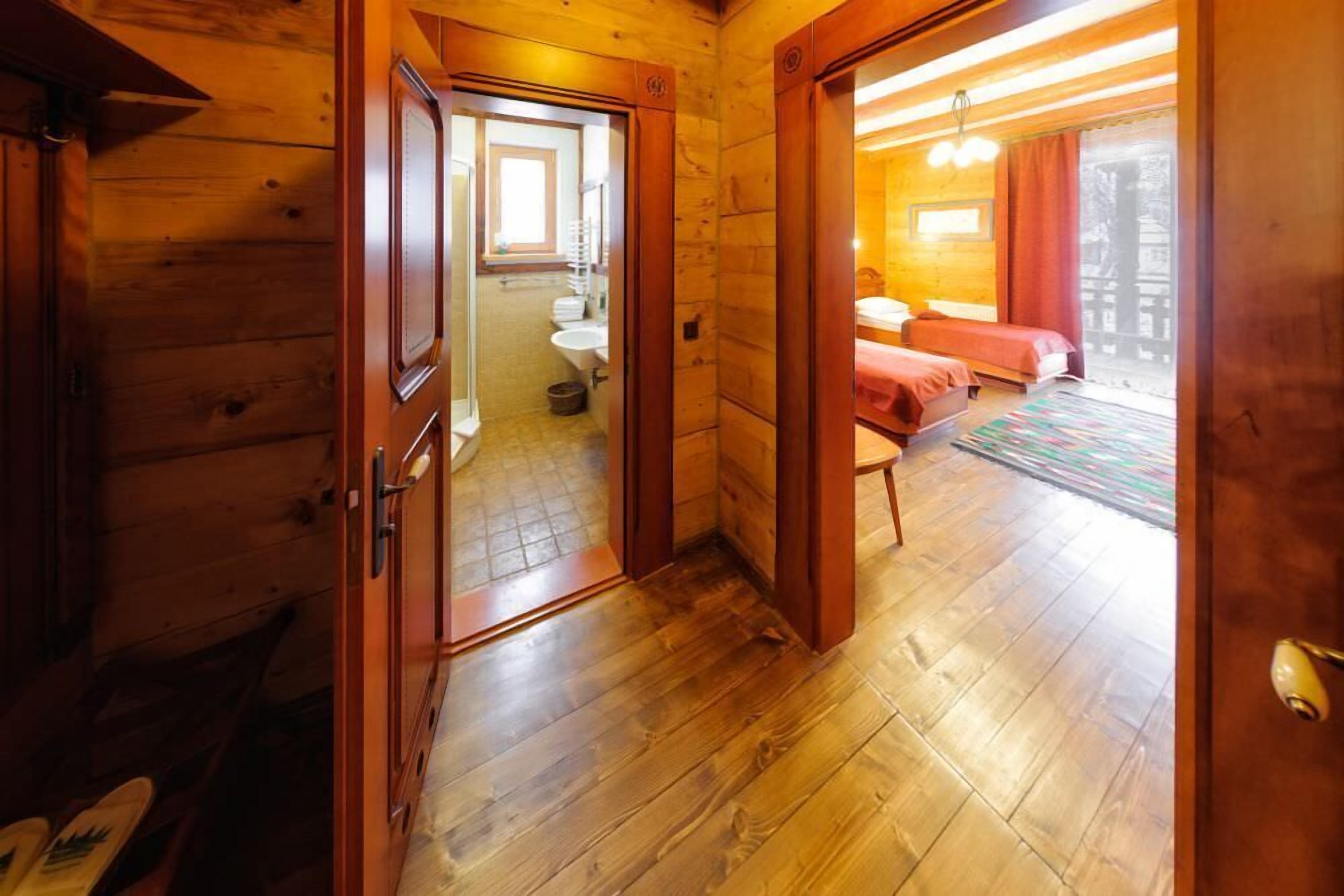 Interior de uma cabana de madeira com um banheiro visível através de uma porta aberta, uma cama com um cobertor vermelho e piso de madeira.