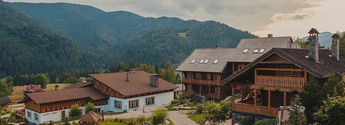 Una casa de madera tradicional con techo inclinado y un balcón, rodeada de abundante vegetación y montañas al fondo.