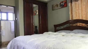 Una cama bien hecha con cabecera de madera, un armario cerrado y un baño visible a través de una puerta entreabierta.
