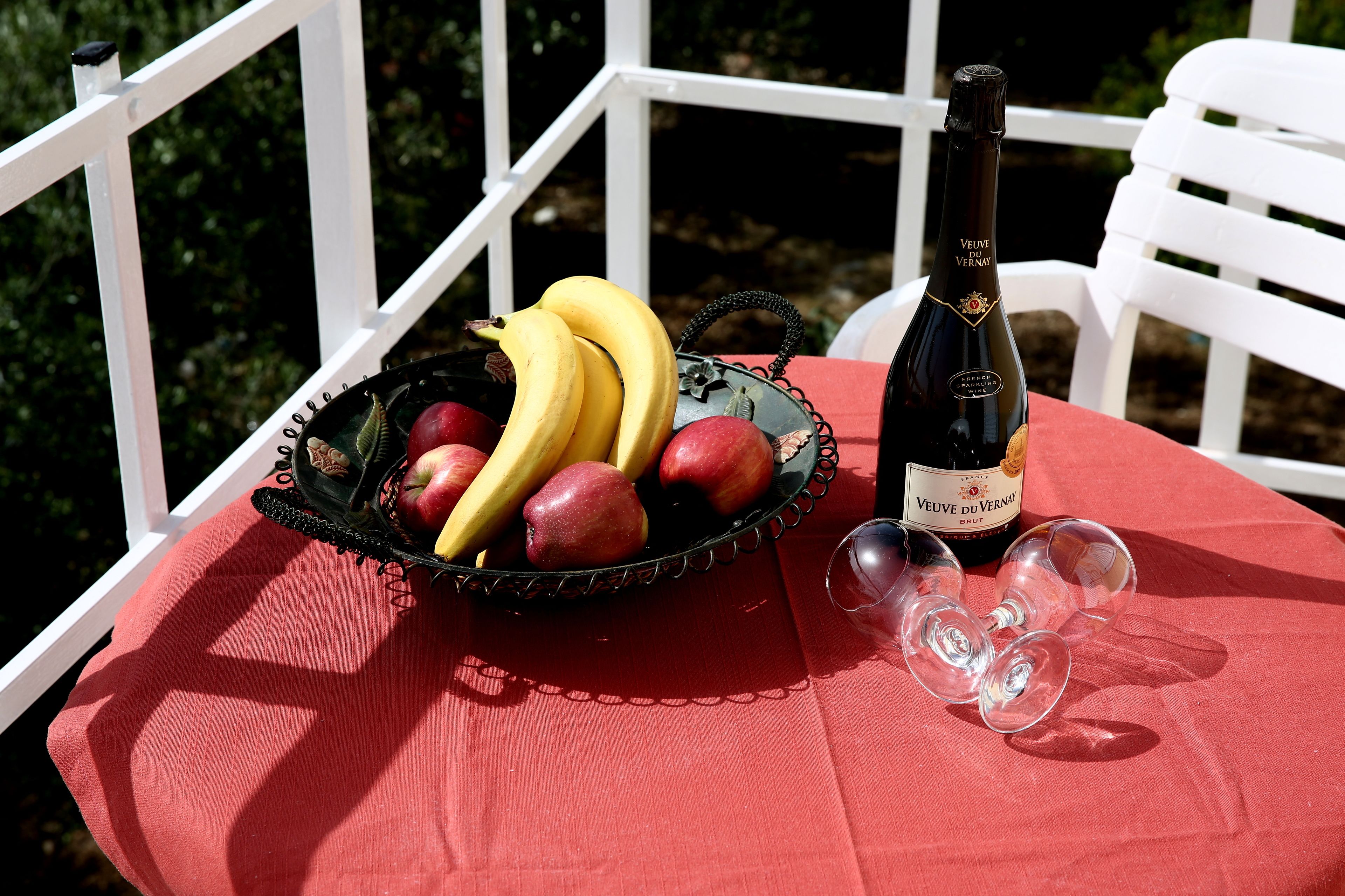 A table with a red tablecloth, a black basket with fruits, a bottle of Veuve du Vernais wine, and two wine glasses.