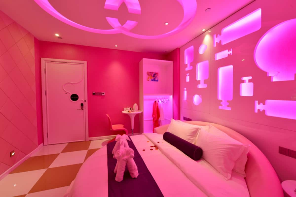 A pink-themed hotel room with a bed, a teddy bear, a small table, and a wall with various icons.