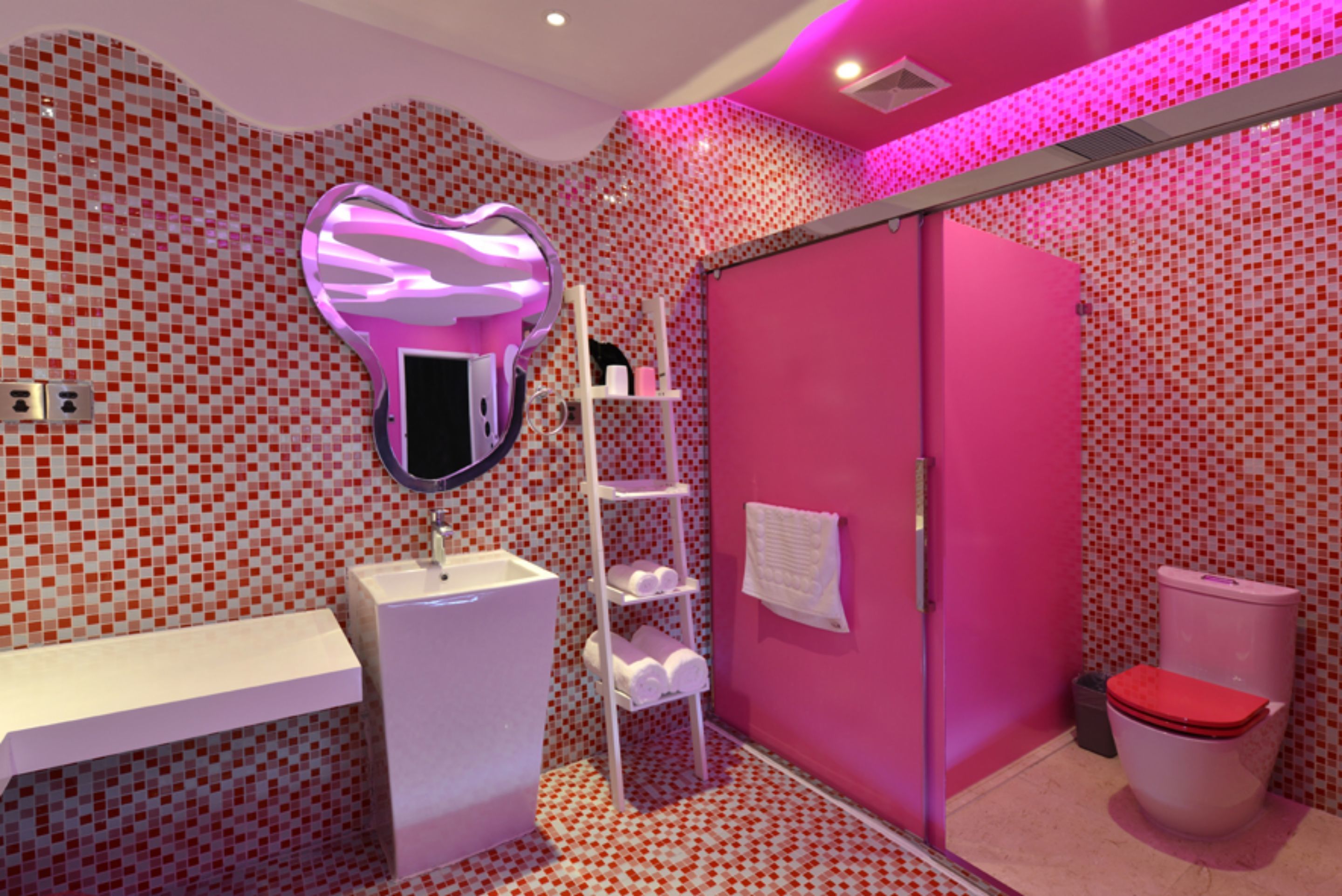 A bathroom with a heart-shaped mirror, a white sink, a pink shower enclosure, and a toilet with a red seat.