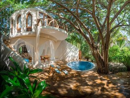 A white, curved building with a pool and outdoor seating area surrounded by trees.