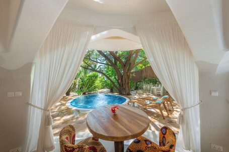 A room with a view of an outdoor swimming pool, a wooden table with chairs, and white curtains framing the window.
