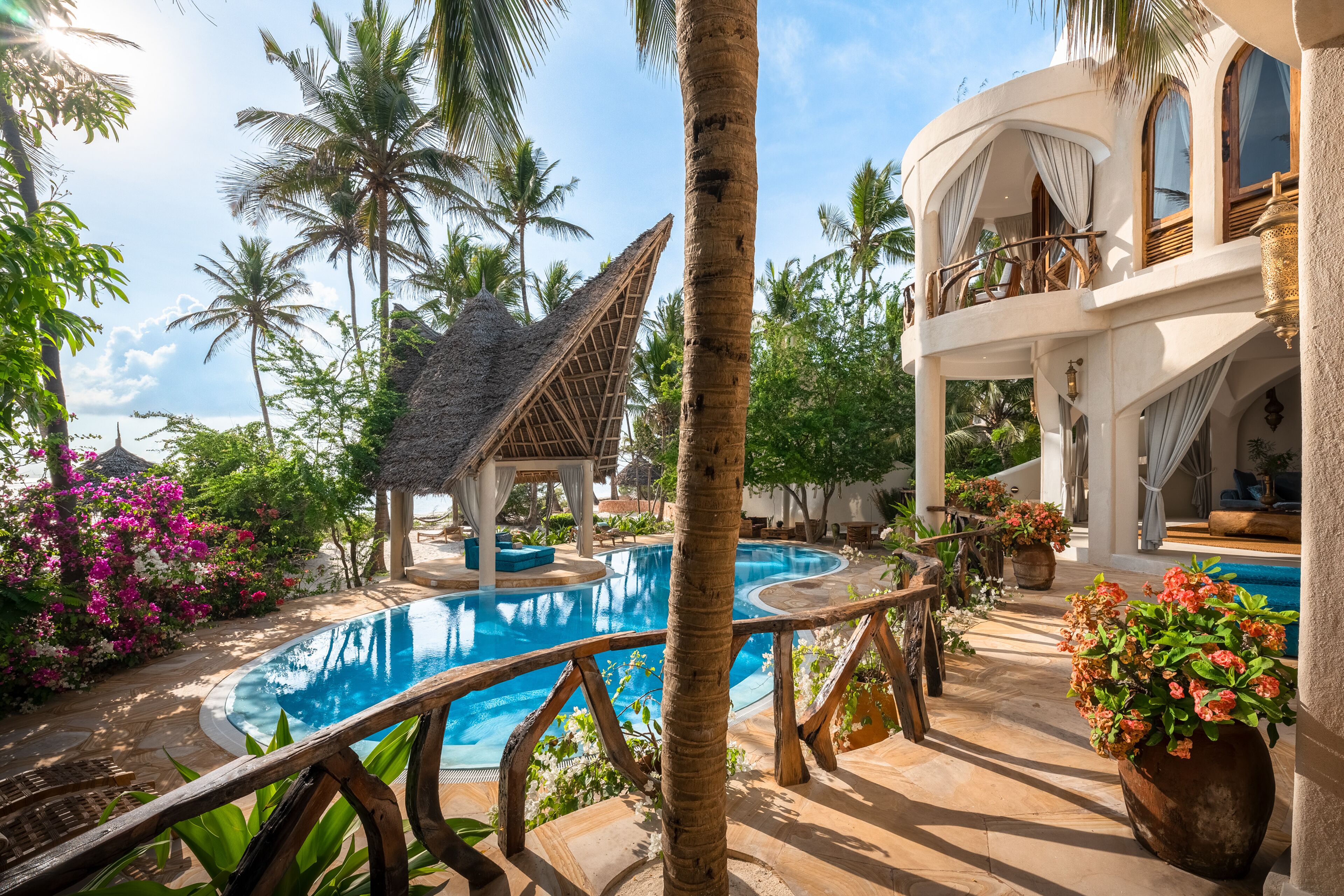 A pool area with a thatched-roof structure, palm trees, and a building with balconies.