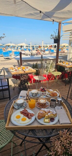 An outdoor breakfast setup with a variety of food items on trays, overlooking a beach with umbrellas and lounge chairs. - Hotel Mirage Bellaria (Bellaria-Igea Marina)