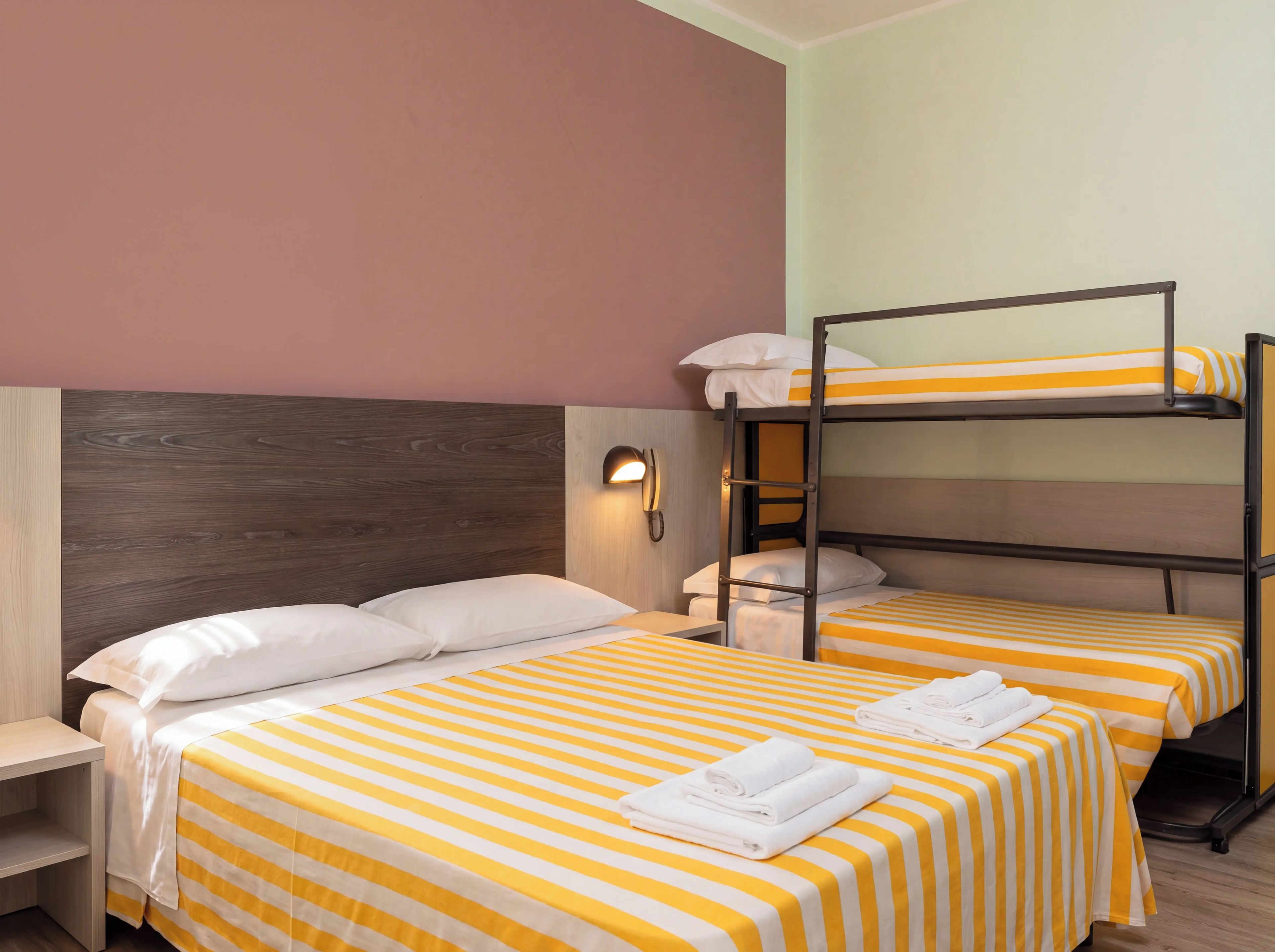 A bunk bed room with two beds, each with a yellow-striped bedspread and white pillows. There is a bedside table with a lamp, and a wall-mounted phone.