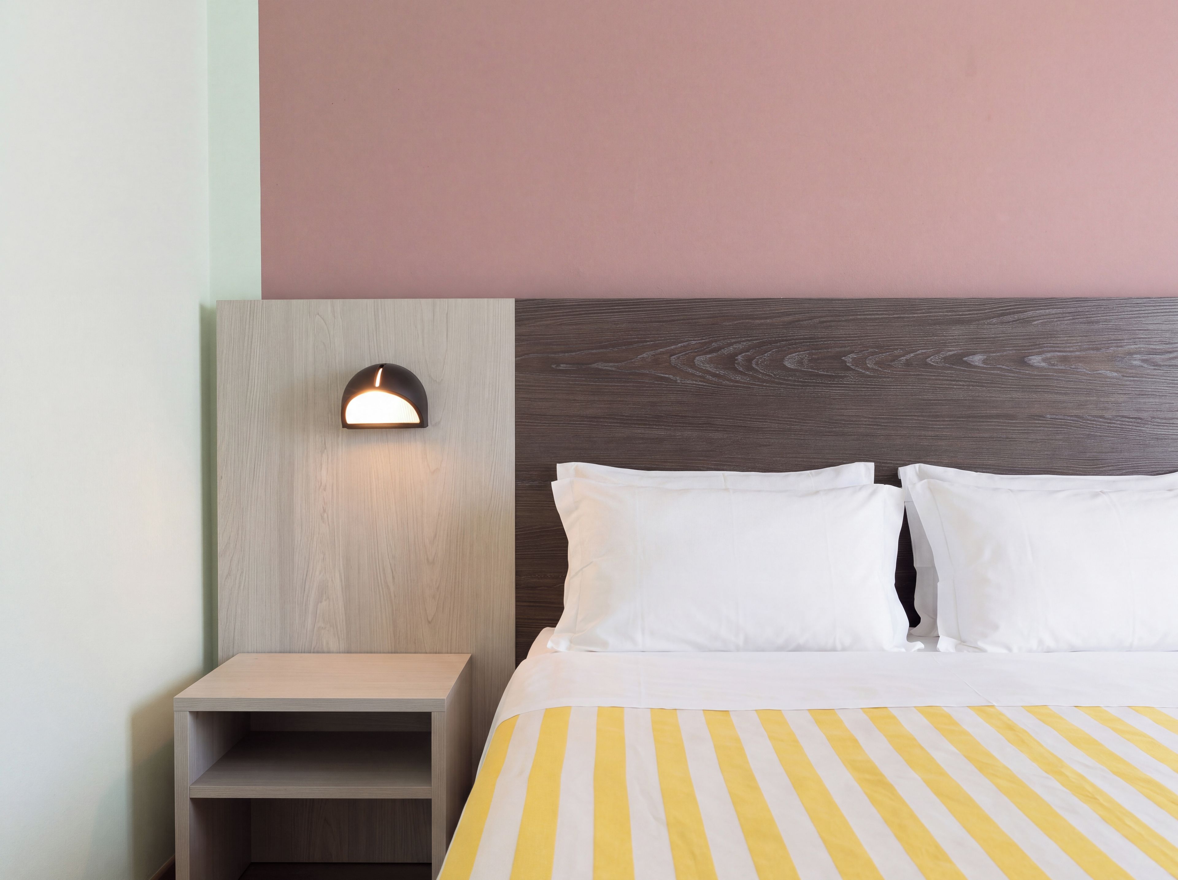 Quarto de hotel com cama, mesa de cabeceira e luminária fixada na parede.