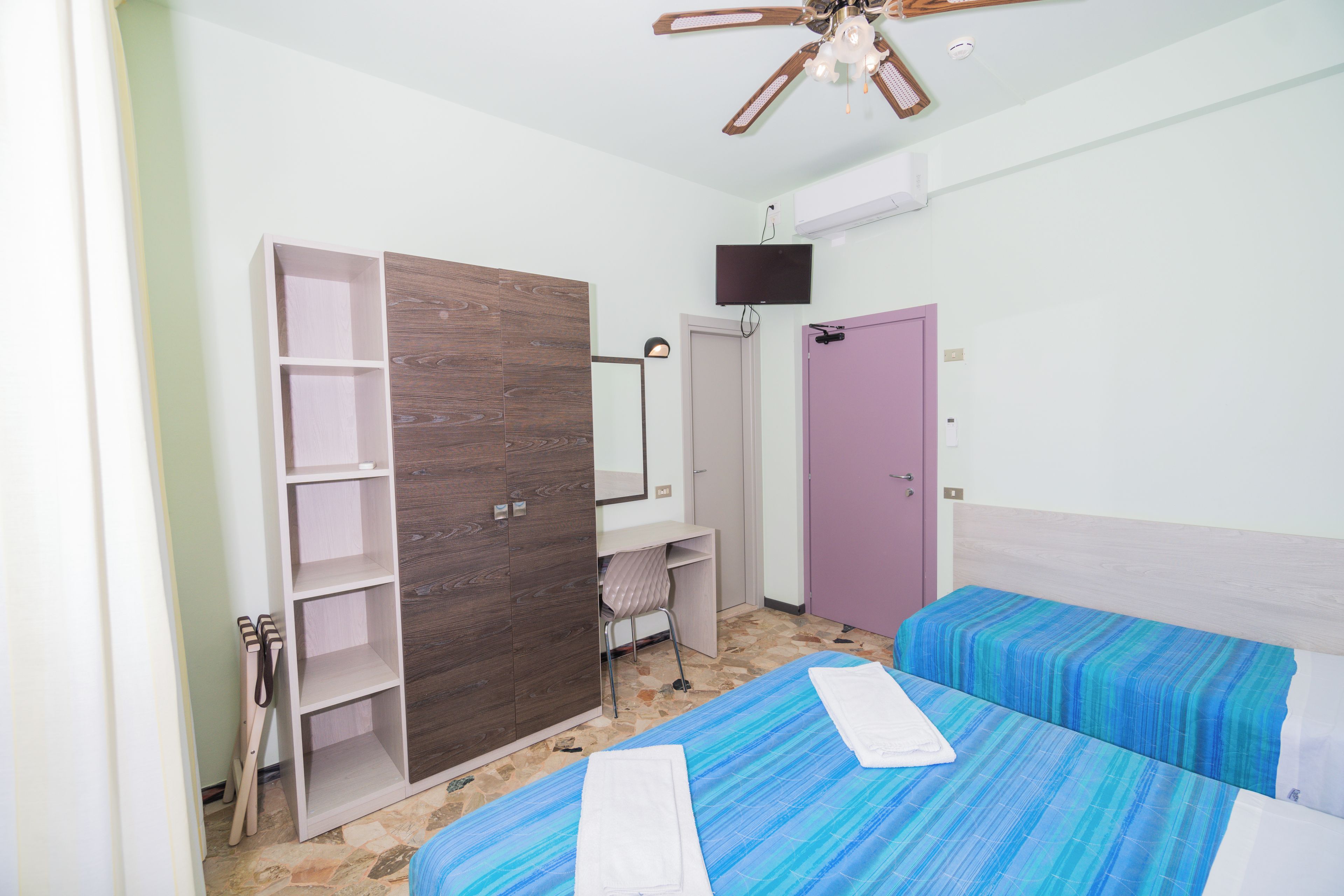A bedroom with two beds, a wardrobe, a desk, a chair, a ceiling fan, and a television.