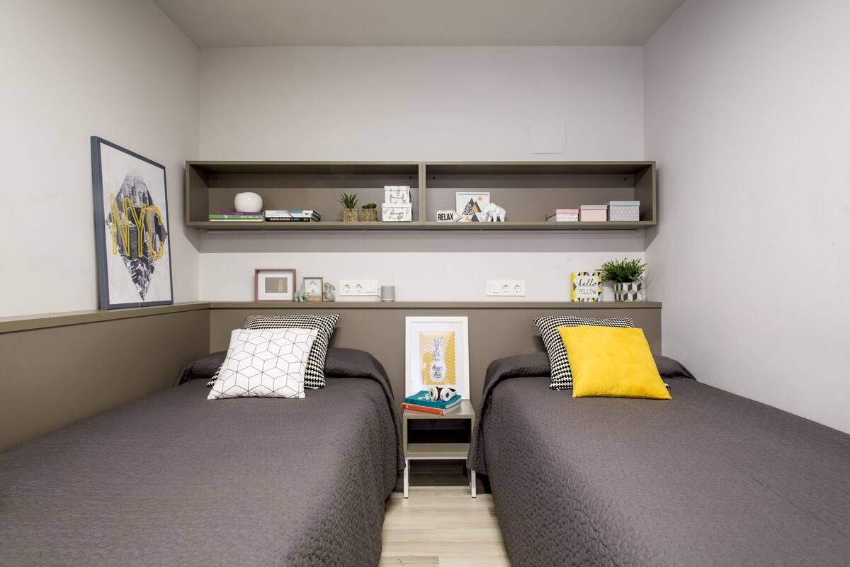 Une chambre double avec deux lits, une étagère avec des livres et des objets décoratifs, et un tableau encadré au mur.