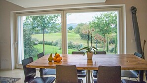 Un comedor con una mesa grande, sillas, una orquídea en maceta y vistas a un paisaje verde a través de amplias ventanas.