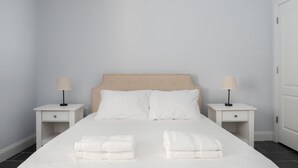A neatly made bed with white linens and two pillows, flanked by two bedside tables with lamps, in a room with light-colored walls and a door on the right. - 6 Bedroom Duplex Near NYC (Jersey City)