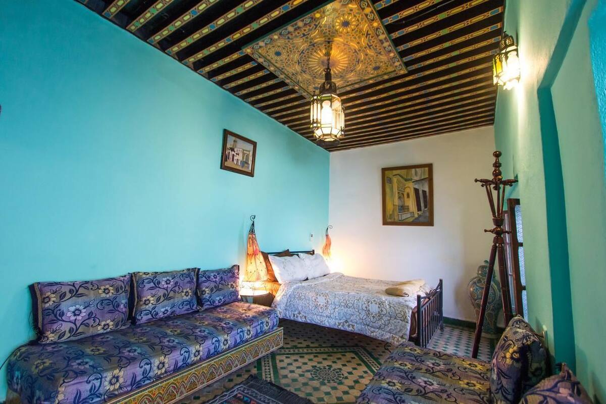 A room with a bed, a sofa, a patterned rug, a decorative ceiling, and wall-mounted lights.