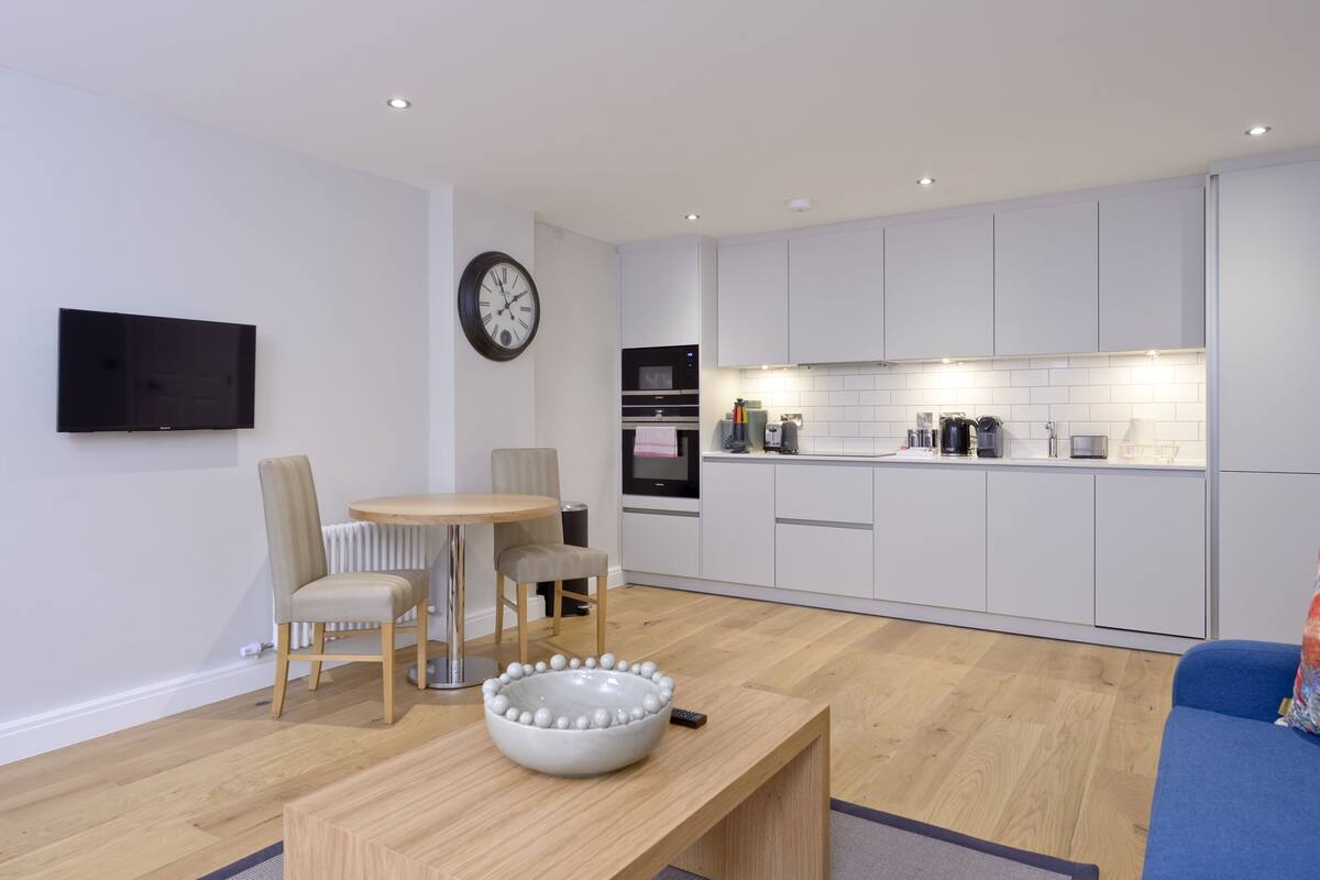 A modern kitchen with white cabinets, a built-in oven, and a microwave. There is a dining area with a round table and chairs.
