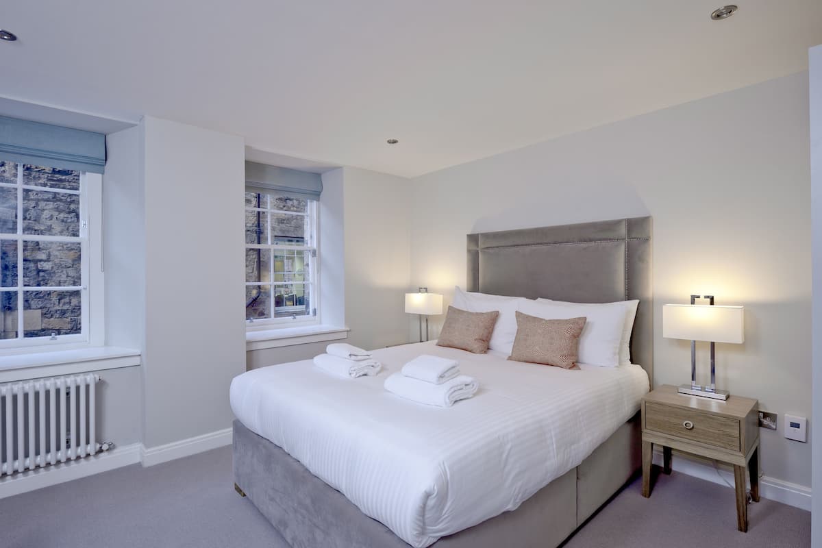 A neatly made bed with white linens and two pillows, a bedside table with a lamp, and a window with a view of a stone wall.