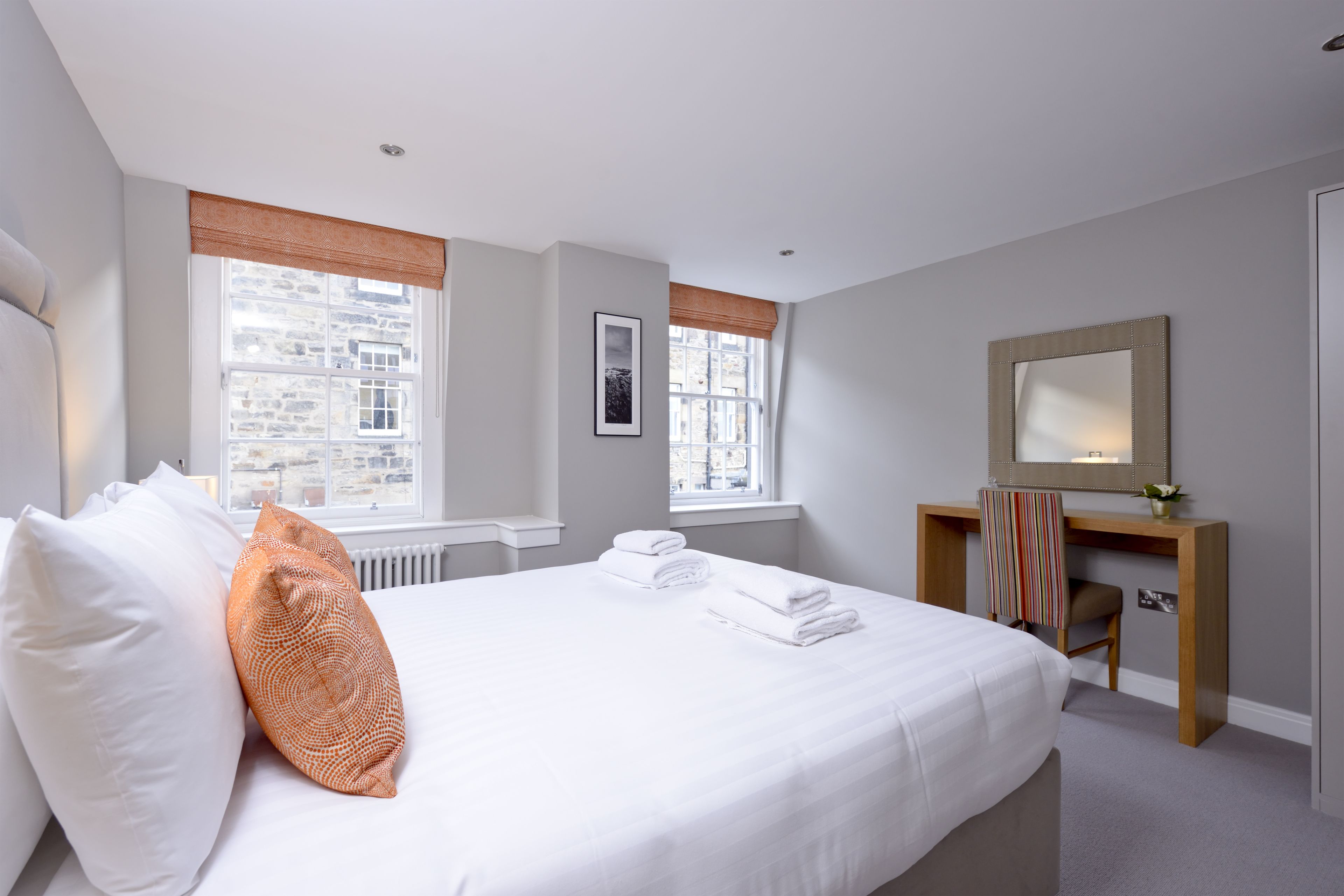 A hotel room with a large bed, two windows, a desk, and a mirror.