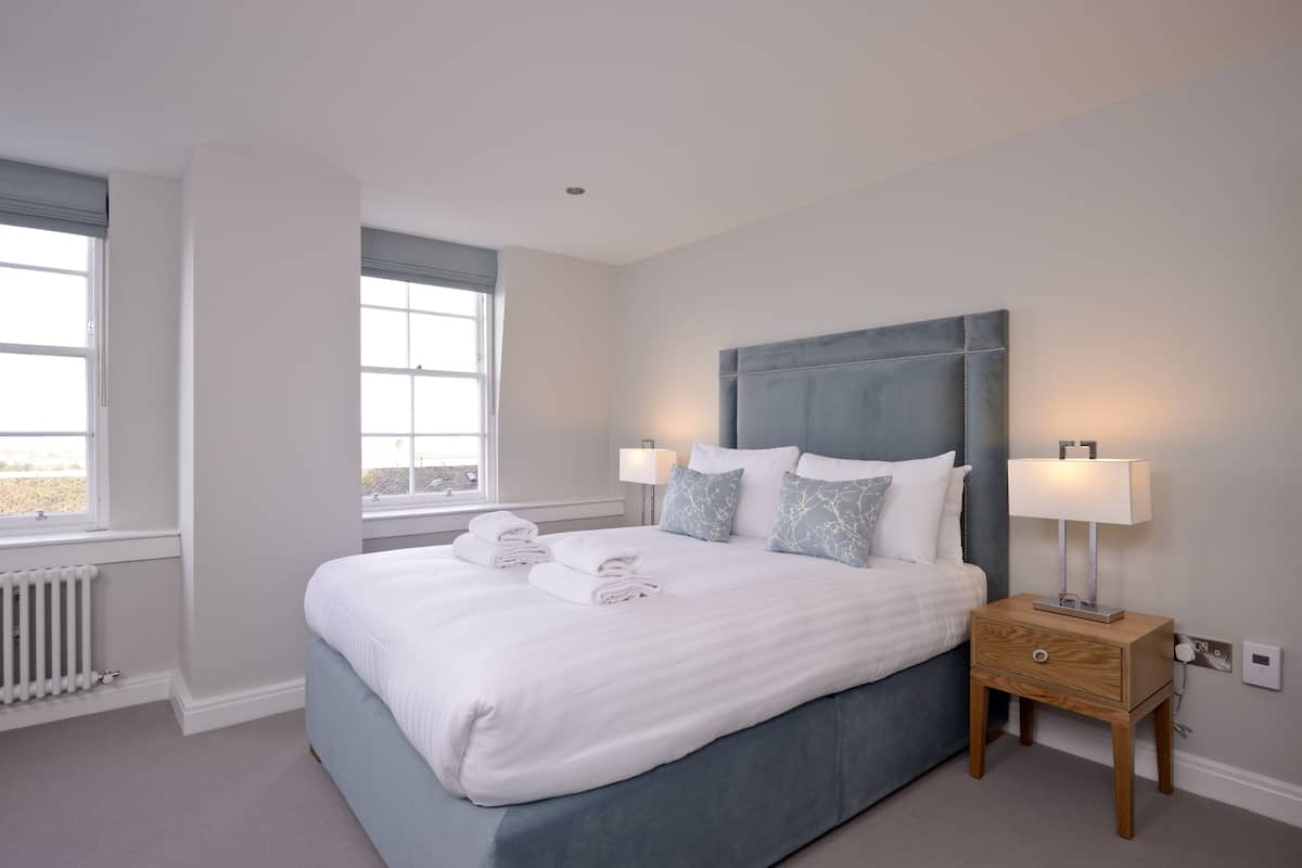 A neatly made bed with white linens and blue bedding, a bedside table with a lamp, and a window with a view.