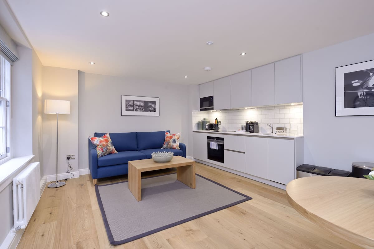 A modern living room with a blue sofa, a wooden coffee table, and a kitchen area with white cabinets and appliances.