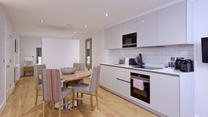 Une cuisine moderne avec des meubles blancs, un four à micro-ondes et un four encastré. On y trouve un coin repas avec une table et des chaises, ainsi qu’une petite table d’appoint avec une lampe.