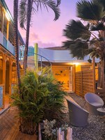 Un espacio exterior tropical con una terraza de madera, palmeras y un área de descanso con una silla y una mesa.
